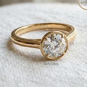 May include: A gold engagement ring with a large, round, clear gemstone. The ring has a detailed band with a beaded design and a bezel setting. The ring is from Sparkora Jewels.
