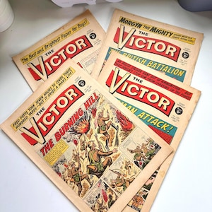 May include: Four vintage copies of "The Victor" comic books are shown. The covers have red and white lettering, with war-themed illustrations. Titles include "The Burning Hill" and "Man Attack!" The comics are dated June 1967.