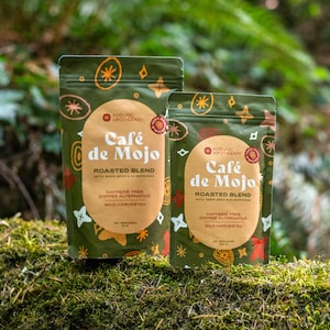 Könnte beinhalten: Zwei grüne Beutel "Café de Mojo" Kaffee-Alternative, mit Text und dekorativen Elementen. Die Beutel sind mit "Roasted Blend" und "Caffeine Free" beschriftet. Der Hintergrund ist eine natürliche Umgebung mit grünem Laub.