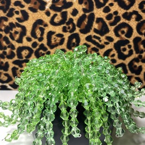May include: A decorative beaded plant in a black pot. The plant is made of light green, faceted beads, with strands cascading down. The background features a leopard print pattern in shades of brown and black.