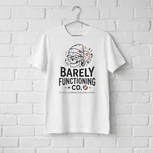 Peut inclure: Un t-shirt blanc à manches courtes avec un motif représentant un crâne portant un bonnet et des lunettes à côté d'une ampoule. Le texte BARELY FUNCTIONING CO. EST. 2023 | ALTERNATIVE & RELATABLE APPAREL est imprimé en noir et orange.