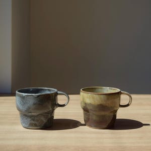 Può includere: Due tazze in ceramica con manici. Una è grigio scuro con una superficie strutturata, l'altra è marrone e gialla. Entrambe le tazze hanno una forma cilindrica e sono su una superficie di legno.