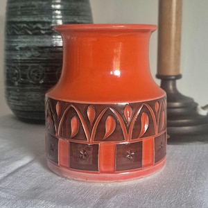 Op de afbeelding: Een keramieken vaas met een feloranje bovenzijde en een onderste band versierd met een donkerbruin geometrisch patroon van bogen en vierkanten. Het oppervlak heeft een glanzende afwerking en bevat kleine, in reliëf aangebrachte bloemmotieven.
