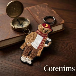 Op de afbeelding: Een geborduurde sleutelhanger van een bruine teddybeer met een rode honkbalpet met een gele letter B, een rode sjaal, een beige jas, een wit shirt, een bruine broek en witte sneakers, terwijl hij een zwarte koffiebeker met witte deksel vasthoudt.