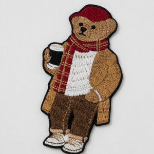 May include: Embroidered patch of a teddy bear wearing a red cap and scarf, brown coat, and pants. The bear is holding a black mug with a white top. The patch is a detailed design with various colors.