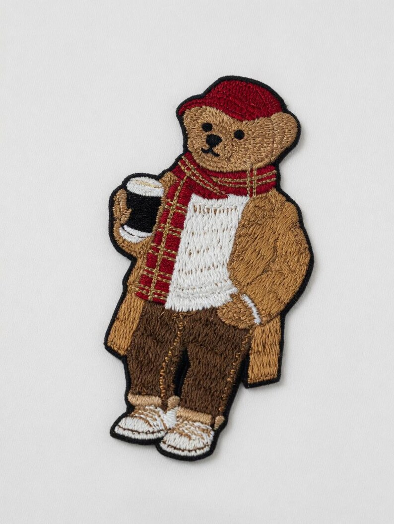 May include: Embroidered patch of a teddy bear wearing a red cap and scarf, brown coat, and pants. The bear is holding a black mug with a white top. The patch is a detailed design with various colors.