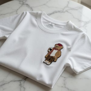 May include: A white t-shirt with a crew neck featuring an embroidered patch of a bear wearing a red hat, scarf, and brown pants, holding a coffee cup. The bear is light brown with white accents. The shirt is on a white marble surface.