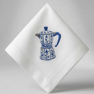 Puede incluir: Una servilleta de tela blanca cuadrada con un bordado central en azul y gris de una cafetera italiana. El diseño muestra una cafetera detallada con motivos florales, un asa azul y un pomo de tapa azul sobre la tela blanca.