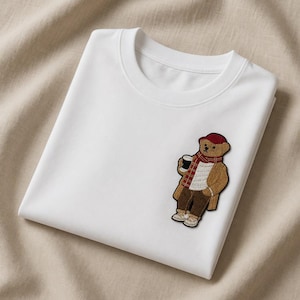 May include: White crew neck t-shirt with a bear patch. The patch features a brown bear wearing a red hat, scarf, and coat, holding a coffee cup. The t-shirt is folded and placed on a beige fabric.