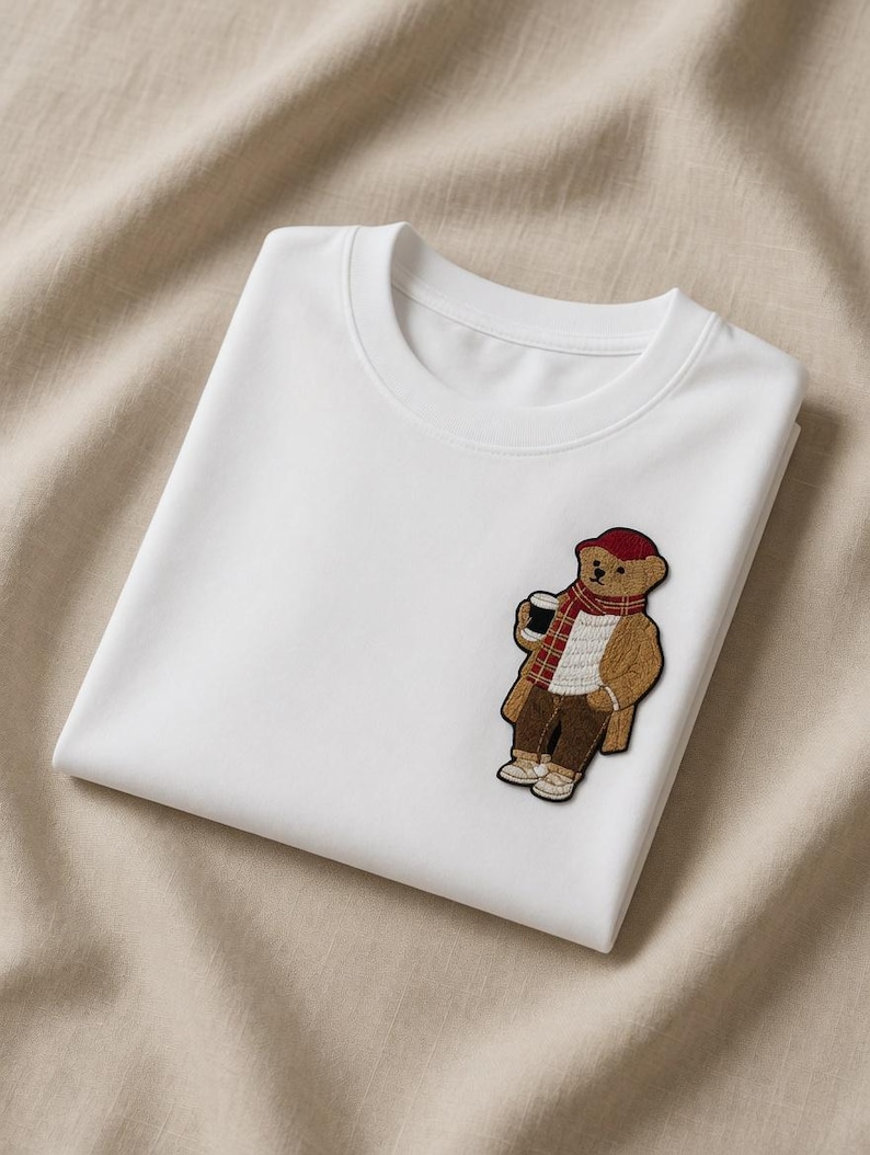 May include: White crew neck t-shirt with a bear patch. The patch features a brown bear wearing a red hat, scarf, and coat, holding a coffee cup. The t-shirt is folded and placed on a beige fabric.