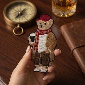 Könnte beinhalten: Ein gestickter Aufnäher, der einen beigen Teddybären mit roter Mütze, rot-gold kariertem Schal, weißem Pullover, beigem Mantel und dunkelbrauner Hose zeigt, während er eine schwarze Kaffeetasse in der Hand hält.