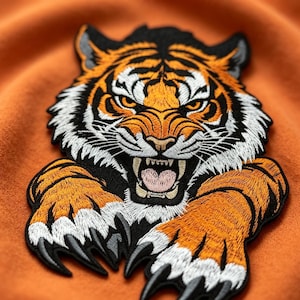 Könnte beinhalten: Ein gestickter Tiger-Aufnäher mit einem fauchenden Tigerkopf in orangefarbenem, schwarzem und weißem Fell, entblößten Zähnen und ausgestreckten Krallen. Das Design ist in detaillierter Stickarbeit mit scharfen Zügen und einem schwarzen Rand ausgeführt.