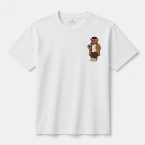 May include: White t-shirt featuring a small embroidered patch of a bear wearing a coat, scarf, and hat, holding a coffee cup. The bear is brown, with a red hat and scarf. The t-shirt has a crew neck and short sleeves.
