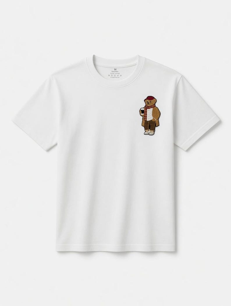 May include: White t-shirt featuring a small embroidered patch of a bear wearing a coat, scarf, and hat, holding a coffee cup. The bear is brown, with a red hat and scarf. The t-shirt has a crew neck and short sleeves.