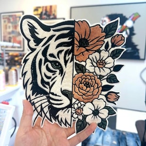 Op de afbeelding: Een geborduurde patch met een gespleten ontwerp. De linkerkant toont een witte tijgerkop met zwarte strepen en een groen oog. De rechterkant toont een bloemstuk met beige en witte bloemen en donkergroene bladeren op een witte achtergrond.