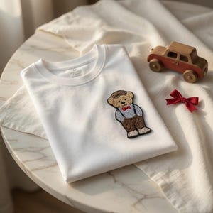 May include: A folded white t-shirt with a teddy bear patch wearing a hat, braces, and a bow tie. A small wooden car and a red bow are next to the shirt on a white surface.