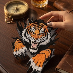 Op de afbeelding: Een geborduurde patch met een woeste tijger met oranje, zwarte en witte vacht, afgebeeld alsof hij door een donker oppervlak breekt met zijn klauwen uitgestrekt en zijn bek open. De tekst 'Coretrims' is zichtbaar in de rechterbenedenhoek.