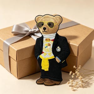 May include: An embroidered patch of a tan bear wearing yellow-rimmed sunglasses, a white turtleneck sweater with a crest, a black blazer, and a yellow sweater tied around the waist, paired with brown shoes, standing against a gift box.