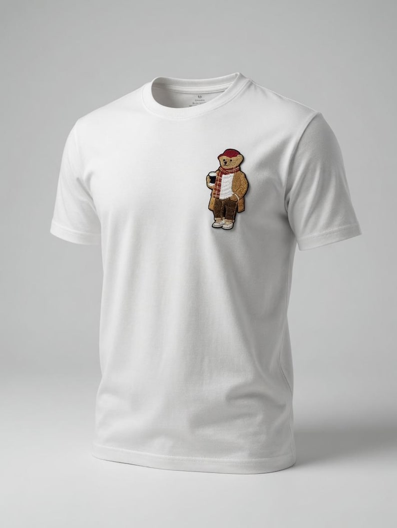 May include: White t-shirt with a small embroidered patch of a bear wearing a red hat, scarf, and coat, holding a coffee cup. The bear is in a standing pose. The shirt has a crew neck and short sleeves.