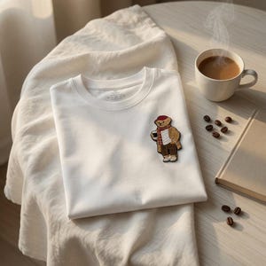May include: A folded white t-shirt with a bear patch, a cup of coffee, and a book on a round table. The bear patch is wearing a hat and scarf. Coffee beans are scattered on the table.