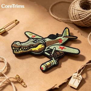 May include: An embroidered patch featuring a cartoon alligator head integrated into the fuselage of a light green military-style aeroplane. The plane has red star insignias on the wings and is depicted carrying a bomb underneath its fuselage.