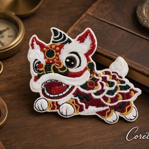May include: An embroidered patch featuring a stylized lion dancer character with white, red, dark green, and gold thread. The design depicts a creature with a wide open mouth, large eyes, and a patterned body, finished with white edges.