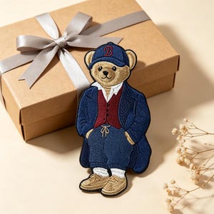 Könnte beinhalten: Ein gestickter Aufnäher mit einem beigen Teddybären, der eine marineblaue Baseballkappe mit einem roten B, ein weißes Hemd, eine dunkelrote Weste, einen marineblauen Mantel, eine marineblaue Hose mit Kordelzug und beige Sneaker trägt.