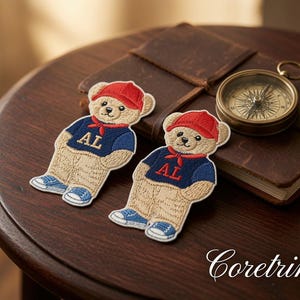 Puede incluir: Dos parches bordados que muestran un oso de peluche color canela con una gorra de béisbol roja, un pañuelo rojo, un suéter azul marino con las letras AL en dorado y zapatillas azules. Los parches presentan detalles de costura intrincados.