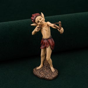 Peut inclure: Figurine d'elfe espiègle, tenant un lance-pierre. L'elfe porte un bonnet et une jupe rouges, et un torse bronzé. Il se tient sur une base marron texturée, avec une expression malicieuse. Le thème général est la fantaisie et le folklore.