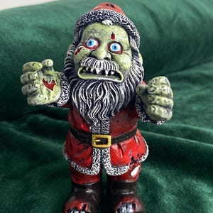 Op de afbeelding: Een zombie-kerstmanfiguur met een bleekgroene huid, felblauwe ogen en een gehavend rood pak. Het figuur heeft een witte baard, een zwarte riem met een gouden gesp en donkere laarzen. De handen zijn uitgestrekt met zichtbare wonden.