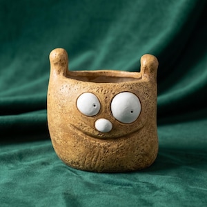 Peut inclure: Un pot de fleurs en céramique beige à la texture granuleuse, représentant une créature avec deux petites oreilles dressées, deux grands yeux blancs circulaires à pupilles noires et un petit nez blanc. Une bouche incurvée est gravée sur le devant.
