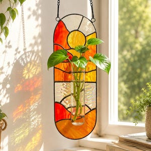 Könnte beinhalten: Ein hängender Glas-Pflanztopf mit einem Sonnenuntergangsdesign in Orange, Gelb und Weiß. Der Pflanztopf enthält ein klares Glasrohr mit grünen Pflanzen. Eine schwarze Kette hängt den Pflanztopf auf.