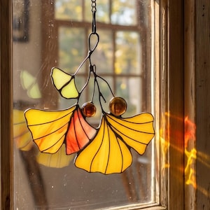 Puede incluir: Atrapasoles de vidrio manchado con hojas de ginkgo en tonos amarillos, naranjas y verdes. El atrapasoles cuelga de una cadena negra y se exhibe en un marco de ventana de madera. Las hojas de ginkgo están detalladas con líneas negras.