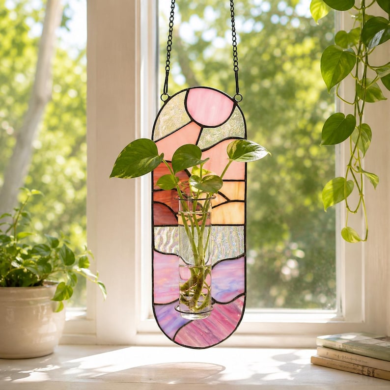 May include: A stained glass window panel in shades of pink, orange, and purple, with a black metal frame and chain for hanging. A small glass vase holds a green plant. The panel is hanging in front of a window.