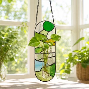 May include: A stained glass plant holder with a clear glass tube for plants. The holder is a vertical oval shape with green, yellow, and blue glass pieces. It hangs from a black chain. The plant has green leaves.