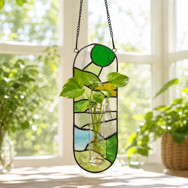 May include: A stained glass plant holder with a clear glass tube for plants. The holder is a vertical oval shape with green, yellow, and blue glass pieces. It hangs from a black chain. The plant has green leaves.