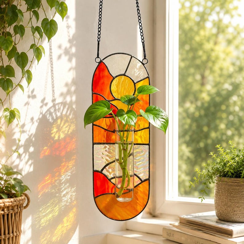 May include: A stained glass hanging planter with a vibrant sunset design in shades of orange, yellow, and white. The planter holds a clear glass tube with green leafy plants. A black chain suspends the planter.