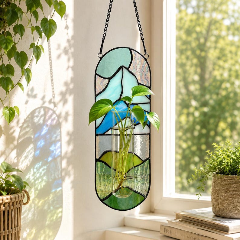 May include: A stained glass hanging planter with a black metal frame. The glass features a mountain scene in shades of blue, green, and white. A clear glass tube holds a green plant with large leaves. The planter hangs from a black chain.