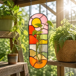 May include: A stained glass window hanging with a landscape design. The window features a yellow sun, pink sky, and colorful terrain. A clear glass vase holds a green plant. The window is suspended by a black chain.
