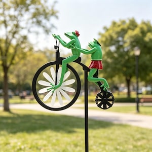 Può includere: Un'originale paletto da giardino con due figurine di rana verde su una bicicletta nera. La ruota anteriore ha pale bianche a forma di margherita e la rana sul retro indossa una gonna rossa. Lo sfondo mostra un parco.