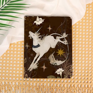 May include: A rectangular notebook featuring a dark brown cover with a white unicorn illustration. The unicorn wears a dark blue collar and is surrounded by white trumpet-shaped flowers, gold star shapes, and swirling vine patterns on a dark background.