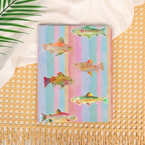 May include: A rectangular notebook featuring a vertical striped pattern in soft pastel shades of blue, pink, and tan. The cover is decorated with five distinct watercolor-style illustrations of trout fish, each displaying varied colors and markings.
