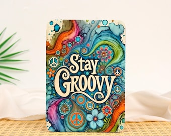 May include: A rectangular sign featuring the text Stay Groovy in a cream-colored script font. The background displays a vibrant, swirling alcohol ink pattern in shades of blue, teal, orange, pink, and purple, accented with small peace signs and flowers.
