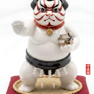 Op de afbeelding: Een witte keramische sumo kat figuur met een zwarte mawashi, rode kraag en bel. De kat heeft traditionele sumo worstelaar gezichtsverf en houdt een miniatuur huis met figuren vast. Het staat op een rood kussen.