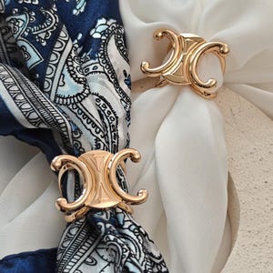 May include: Two gold-toned metal scarf rings featuring a symmetrical, stylized double-C design. One ring secures a navy blue and white paisley patterned scarf, while the other holds a plain white scarf, showcasing the polished metallic finish.