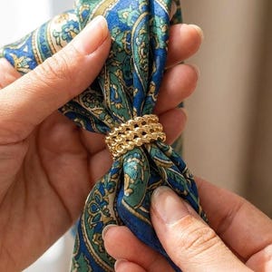 May include: A gold-toned metal scarf ring featuring a triple-layered twisted rope design, securing a blue and green patterned silk scarf. The accessory has a polished finish and a cylindrical shape that holds the fabric in a gathered knot.
