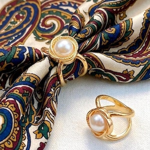 May include: Two gold-toned metal rings featuring a central white pearl set within a textured, circular frame. One ring is shown securing a silk scarf with a multi-colored paisley pattern in shades of blue, green, red, and cream, while the other sits nearby.