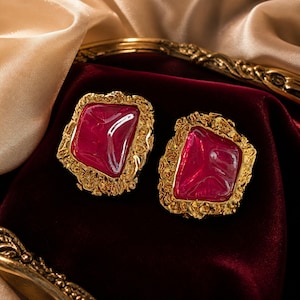 May include: A pair of earrings featuring irregular, deep pink translucent stones set within textured, gold-toned metal frames. The metalwork has a rough, organic appearance surrounding the smooth, glossy, and slightly uneven surfaces of the pink stones.