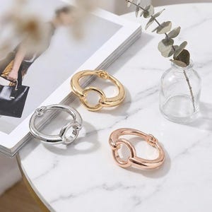 May include: Three metal rings in gold, silver, and rose gold tones. Each ring features a thick, polished band with a smaller circular link attached to the main body, creating a connected, interlocking design with a smooth, reflective finish.