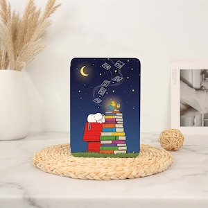 Op de afbeelding: Een rechthoekige beschermhoes met een nachtelijke illustratie van Snoopy die op een rood hondenhok ligt naast een hoge stapel kleurrijke boeken. Een kleine gele vogel zit bovenop de boeken onder een maansikkel en een sterrenhemel.